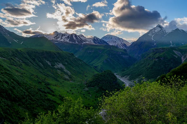 Koyu yeşil vadinin üzerindeki altın gün batımı ışığı ve parlayan bulutların altındaki uzak kar zirveleri.
