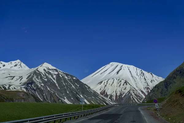 Boş asfalt karayolu, sarp çimenli yamaçların arasına tırmanıyor. Derin mavi gökyüzünün altındaki beyaz karla kaplı dağlara doğru.