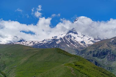 Gürcistan 'daki ikonik Kazbek Dağı parlak mavi gökyüzünün altındaki yemyeşil tepelerin üzerinde yükseliyor.