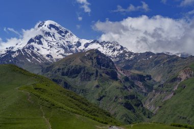 Gürcistan 'daki ikonik Kazbek Dağı parlak mavi gökyüzünün altındaki yemyeşil tepelerin üzerinde yükseliyor.