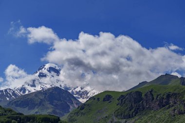 Gürcistan 'daki ikonik Kazbek Dağı parlak mavi gökyüzünün altındaki yemyeşil tepelerin üzerinde yükseliyor.