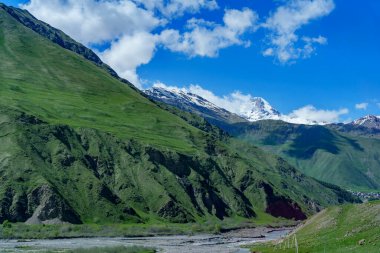 Uçsuz bucaksız çimenli yamaçlar, geniş bir nehir yatağının üzerinde yükseliyor. Parlak bir gökyüzünün altında karlı tepeler var..