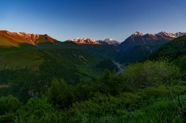 Karlı dağlarda hafif alacakaranlık ışığı parlarken derin yeşil vadide bir nehir esiyor..