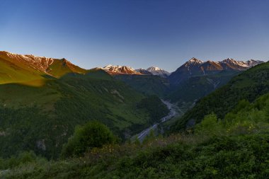 Karlı dağlarda hafif alacakaranlık ışığı parlarken derin yeşil vadide bir nehir esiyor..