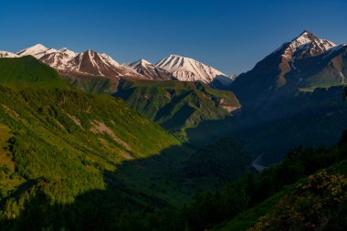 Parlak sabah güneşi karlı Kafkasya tepelerini aydınlatırken gölgeli yeşil bir vadi aşağıda uzanır..