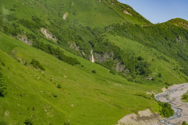 İnce bir şelale taşlı bir nehir ve çayırın üzerindeki yeşil bir vadiye düşüyor..