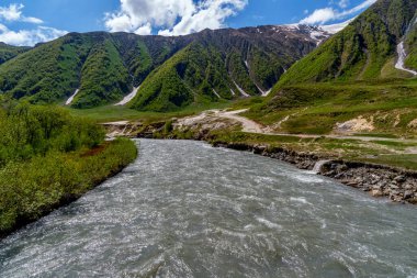 Geniş gri bir nehir sarp yeşil yamaçların ve bulutlarla dolu gökyüzünün altındaki vadide kıvrılır..