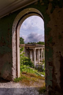 Dıştaki çökmüş iç kemer çerçeveleri aşırı gelişmiş klasik sütunlar, soyulmuş duvarlar, yemyeşil bitki örtüsü ve bulutlu gökyüzü