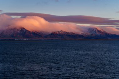 Karla kaplı dağlar pembe alpenglasla yıkanmış alacakaranlıkta sakin denizin üzerine yansıyor.