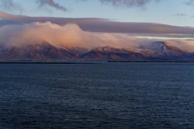 Geniş karlı dağ zinciri, gün batımında denizin üzerinde, katmanlı sislerin arasında pembe parlar.