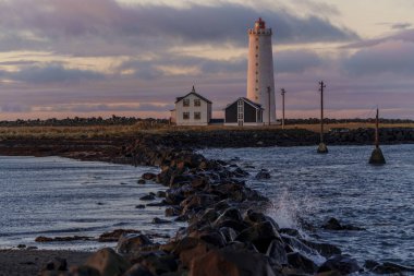 Beyaz çizgili deniz feneri ve dalgalı su üzerindeki evler okyanusun üzerinde pembe günbatımı ışığıyla yıkanmış.