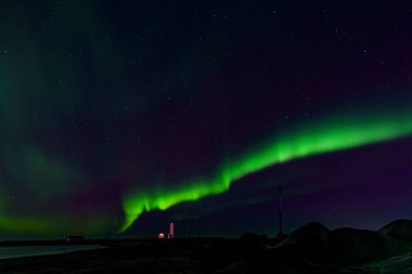 Canlı yeşil Kuzey Işıkları, yıldızlı karanlık gökyüzünde parıldayan deniz feneri, küçük kulübe, kıyı ön planında kayalar üzerinde kavis oluşturur..