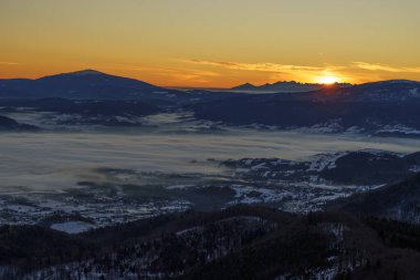 Alçak bulutlar vadiyi karla kaplı tepeler ve aşağıdaki yerleşim yerleriyle doldururken güneş tepeden bakıyor..