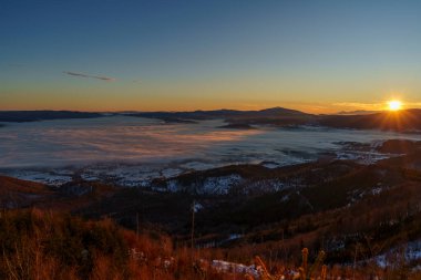 Ufuktaki parlak güneş, kışın karlı tepelerin ve ormanlık tepelerin üzerinde bir bulut denizi aydınlatır..