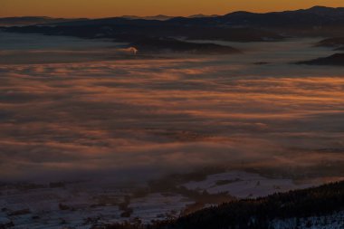 Ilık şafak boyaları karlı vadinin üstünde yuvarlanan sisleri, ufukta karanlık dağ siluetleri.