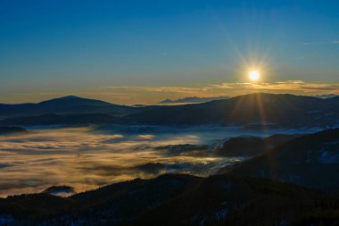 Alçak bulutlar ve sis karlı vadileri doldururken parlak güneş, katmanlı dağların tepelerinde ışıldıyor..