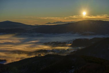 Uzak tepelerin üzerindeki parlak güneş, alçak bulutları ve sisi aydınlatır. Katmanlı çıkıntıları ve aşağıdaki karlı manzarası vardır..