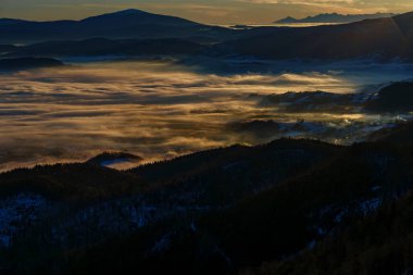 Yumuşak gündoğumu ışığı sislerin içinde parlayan parçalar yaratır. Karanlık orman yamaçları ve uzak dağları vardır..