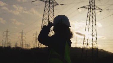 Genç İnşaatçı Kadın, yavaş çekim sahnesiyle ekranda zıplıyor. Elektrik kuleleriyle sahada. Günbatımı arka planında yüksek voltajlı elektrik pilonu olan elektrik mühendisi. çalışma konsepti.