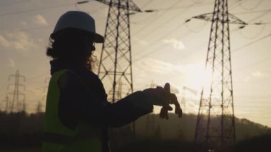 Genç İnşaatçı kadın eldivenlerini yavaş çekim sahnesiyle temizliyor. Elektrik kuleleriyle sahada. Günbatımı arka planında yüksek voltajlı elektrik pilonu olan elektrik mühendisi. çalışma konsepti.
