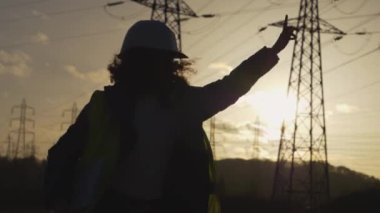 Genç kadın mühendisin silueti, elektrik kuleleriyle birlikte, yavaş çekim sahnesinde mutlu bir şekilde zıplıyor. Günbatımı arka planında yüksek voltajlı elektrik pilonu olan elektrik mühendisi. çalışma konsepti.