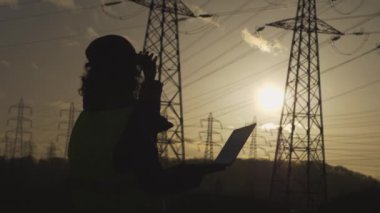 Genç kadın mühendisin silueti ayakta duruyor ve bilgisayarda denetleniyor. Elektrik kuleleriyle birlikte sahada. Günbatımı arka planında yüksek voltajlı elektrik pilonu olan elektrik mühendisi. çalışma konsepti.