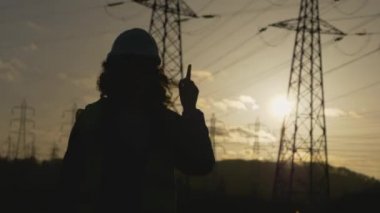 Genç Kadın İnşaatçı 'nın parmağı ve elinin silueti. Elektrik kuleleriyle sahada dur. Günbatımı arka planında yüksek voltajlı elektrik pilonu olan elektrik mühendisi. çalışma konsepti.