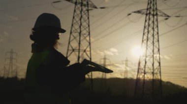 Genç kadın mühendisin silueti bilgisayarı açıp daktilo ediyor. Elektrik kuleleriyle sahada dur. Günbatımı arka planında yüksek voltajlı elektrik pilonu olan elektrik mühendisi. çalışma konsepti.