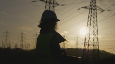 Genç kadın mühendisin silueti bilgisayarı açıp daktilo ediyor. Elektrik kuleleriyle sahada dur. Günbatımı arka planında yüksek voltajlı elektrik pilonu olan elektrik mühendisi. çalışma konsepti.