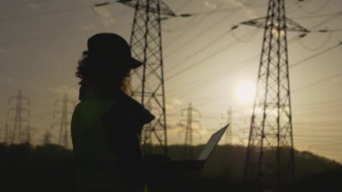 Genç kadın mühendisin silueti ayakta duruyor ve bilgisayarda denetleniyor. Elektrik kuleleriyle birlikte sahada. Günbatımı arka planında yüksek voltajlı elektrik pilonu olan elektrik mühendisi. çalışma konsepti.