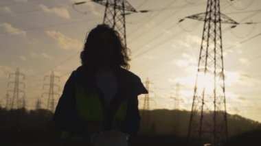 Kıvırcık saçlı genç kadın müteahhit miğferini komik bir şekle sokmuş. Yavaş çekim. Günbatımında yüksek voltajlı elektrik pilonu olan elektrik mühendisi..