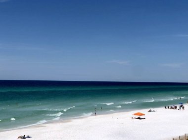 Açık yeşil su ve turuncu çadırı olan güneşli bir tatil plajı manzarası.