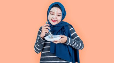 Asyalı tesettürlü kadın evde basit bir yüz maskesi yapmak için çamurla suyu karıştırıyor. Güzellik, cilt bakımı konseptini gerektirir. Turuncu arka planda izole. boş telif alanı