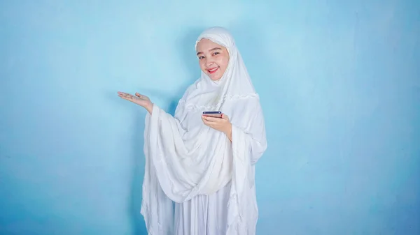 魅力的なアジア系イスラム教徒の女性がスマートフォンを提示して持っています 青い背景に隔離されたフリーコピースペース — ストック写真