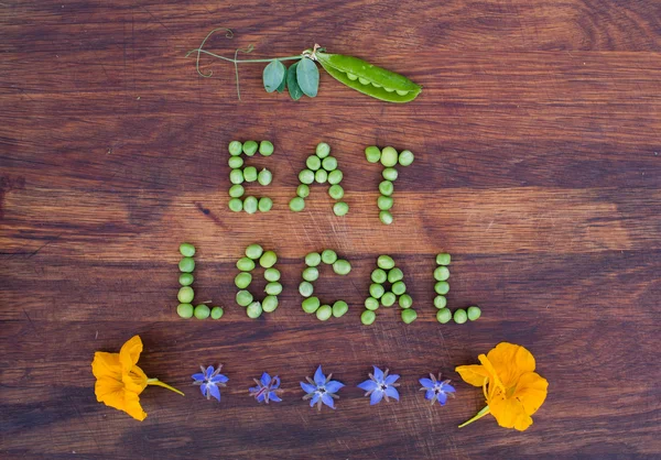 Panneau "Mangez local" fait de pois verts et gousse de pois avec des feuilles sur fond en bois. Fleurs comestibles dans le fond . — Photo