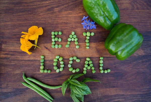 "Mangiare locale "frase fatta di piselli verdi su sfondo di legno. Fiori commestibili, peperoni, basilico e fagiolini su un lato . — Foto Stock