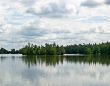 Bir yaz günü kümülüs bulutlarıyla orman, göl ve gökyüzü manzarası.
