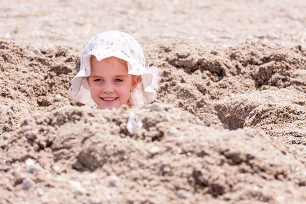 Little girl playing hide and seek hidden in a hole on the beach with only her head sticking out, room for text — Stock Photo, Image