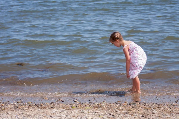 Kleines Mädchen in einem langen Kleid auf der Suche nach Muscheln und Felsen im Meer — Stockfoto