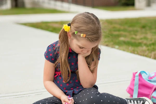 Very sad caucasian little girl sitting on a sidewalk in front of school and worrying or crying — Stock Photo, Image