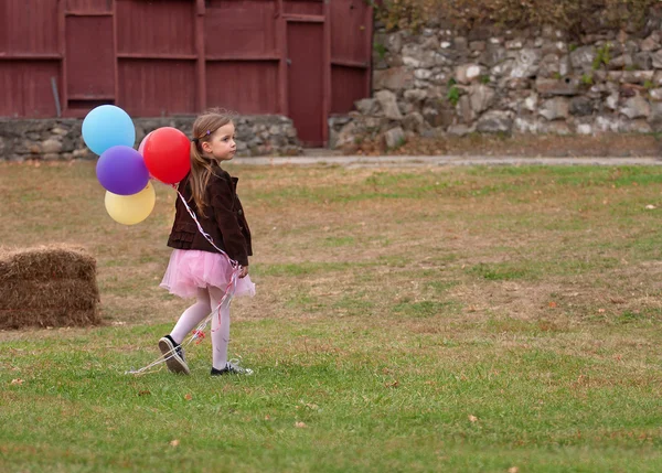 Little girl with a bunch of baloons walking alone on a grass field in the country — Stock Photo, Image