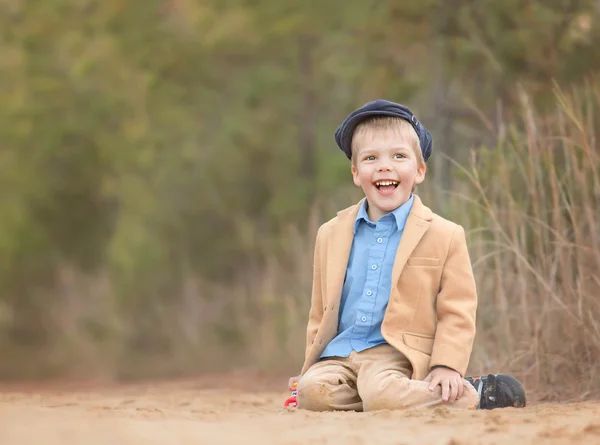 A blond toddler boy sitting on a sand and laughing — Stock Photo, Image