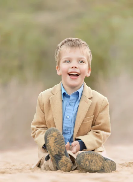 Happy little boy wearing a yellow jacket sittingon a sandy path and laughing — Stock Photo, Image