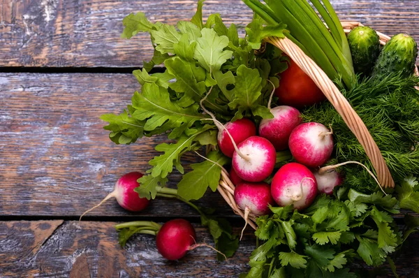 Verduras de primavera y hierbas en una cesta: rábanos, cebollas, perejil, pepino sobre un fondo de madera . — Foto de Stock