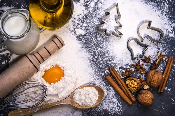 Ingredients for Christmas cookies. Flour, plunger, corolla, wooden spoon, oil, milk, eggs, walnuts on black board — Stock Photo, Image