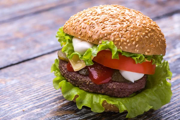 Hamburger mit Rinderschnitzel, frischem Gemüse: Tomaten, Zwiebeln, Salat, Sauce und Semmel mit Sesam auf einem Holzboden — Stockfoto