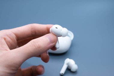 Parmaklar, beyaz kablosuz kulaklığı gri arkaplandaki açık beyaz şarj kutusundan dikkatlice çıkarıyor.