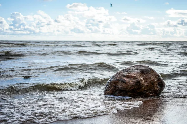 Dalga, deniz kıyısında büyük bir taşa vuruyor. Açık bir gün, açık bulutlar.