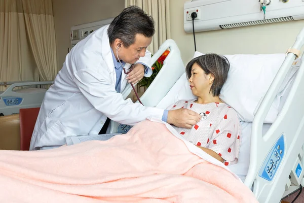 Asian medical doctor examining female patient with stethoscope whom is seated in bed in hospital