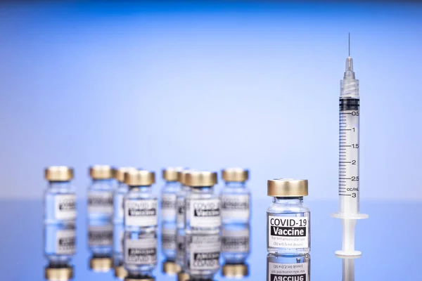 Fokus pada satu botol vaksin Covid-19 dengan jarum suntik, dengan beberapa botol kabur dengan latar belakang biru — Stok Foto